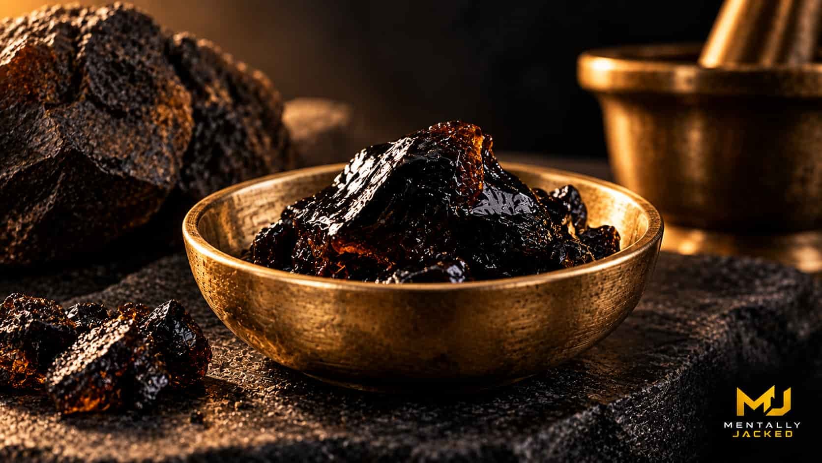 A small piece of dark glossy black resin resting in a plain brass dish on a charcoal surface, representing purified shilajit